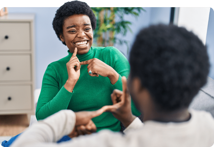 Two people communicate using sign language while seated indoors. One person in a green sweater signs facing the other person, whose hands are visible in the foreground, during a conversation in a bright room.