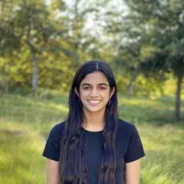 Person with long dark hair, standing outdoors and facing the camera. The person is wearing a dark short-sleeve top and is centered in the frame. The background is softly blurred, featuring green grass, trees, and natural vegetation.