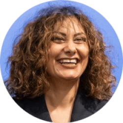 Headshot of a person with shoulder-length curly hair, wearing a dark blazer, against a light blue background.