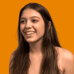 Head-and-shoulders portrait of a person with long dark hair, with a solid bright orange background. The person is turned slightly to one side and is wearing a sleeveless light-colored top. 