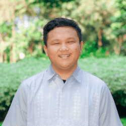 Professional portrait of a person wearing a light-colored button-up shirt, standing outdoors and facing the camera. The background is softly blurred and features green vegetation and trees.
