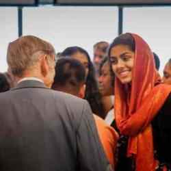 A person wearing a bright orange headscarf and dark clothing stands among a group of people at an indoor event. The person is positioned on the right side of the frame and is engaged in conversation with another attendee whose back is facing the camera. Several other people are visible in the background, which is softly blurred.