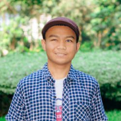 A person wearing a blue-and-white checkered button-up shirt and a dark cap, standing outdoors and facing the camera. The person is centered in the frame, with green shrubs and trees softly blurred in the background.