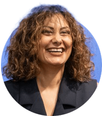 Headshot of a person with shoulder-length curly hair, wearing a dark blazer, against a light blue background.