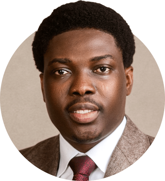 Headshot of a person wearing a brown textured suit jacket, white dress shirt, and red tie, centered against a neutral light background. The image is tightly cropped to the face and upper shoulders and presented in a circular format.